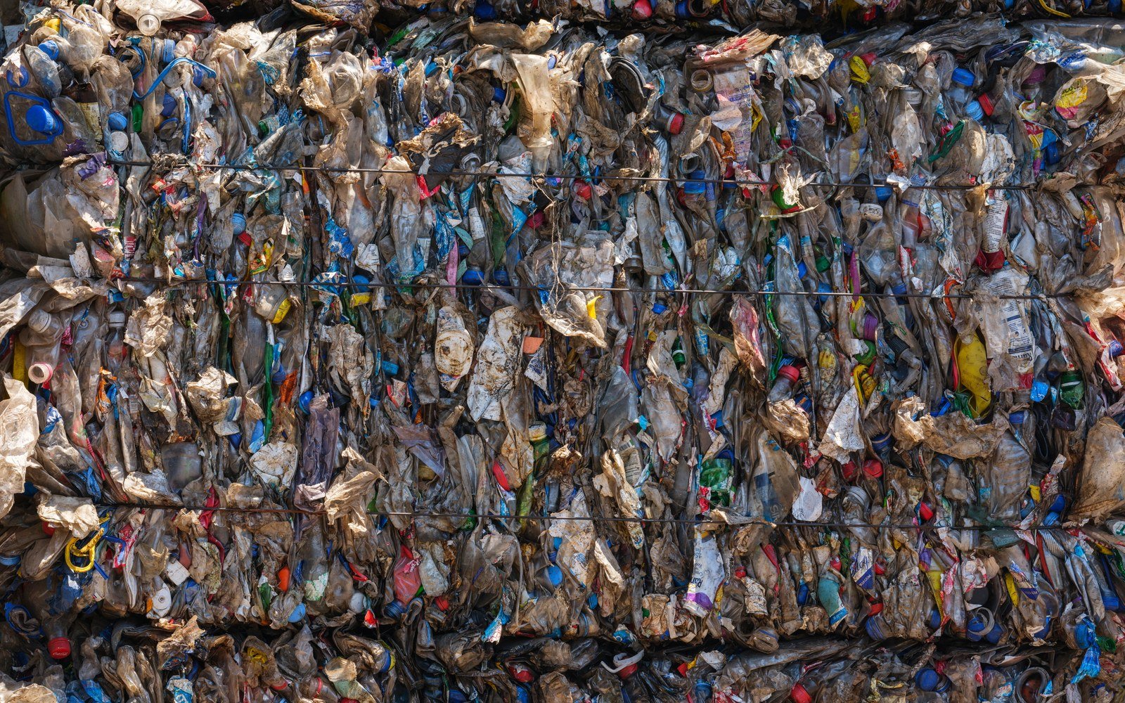 A wall of baled plastic waste held behind horizontal wires at a recycling site, compressed bottles and film visible through the bales