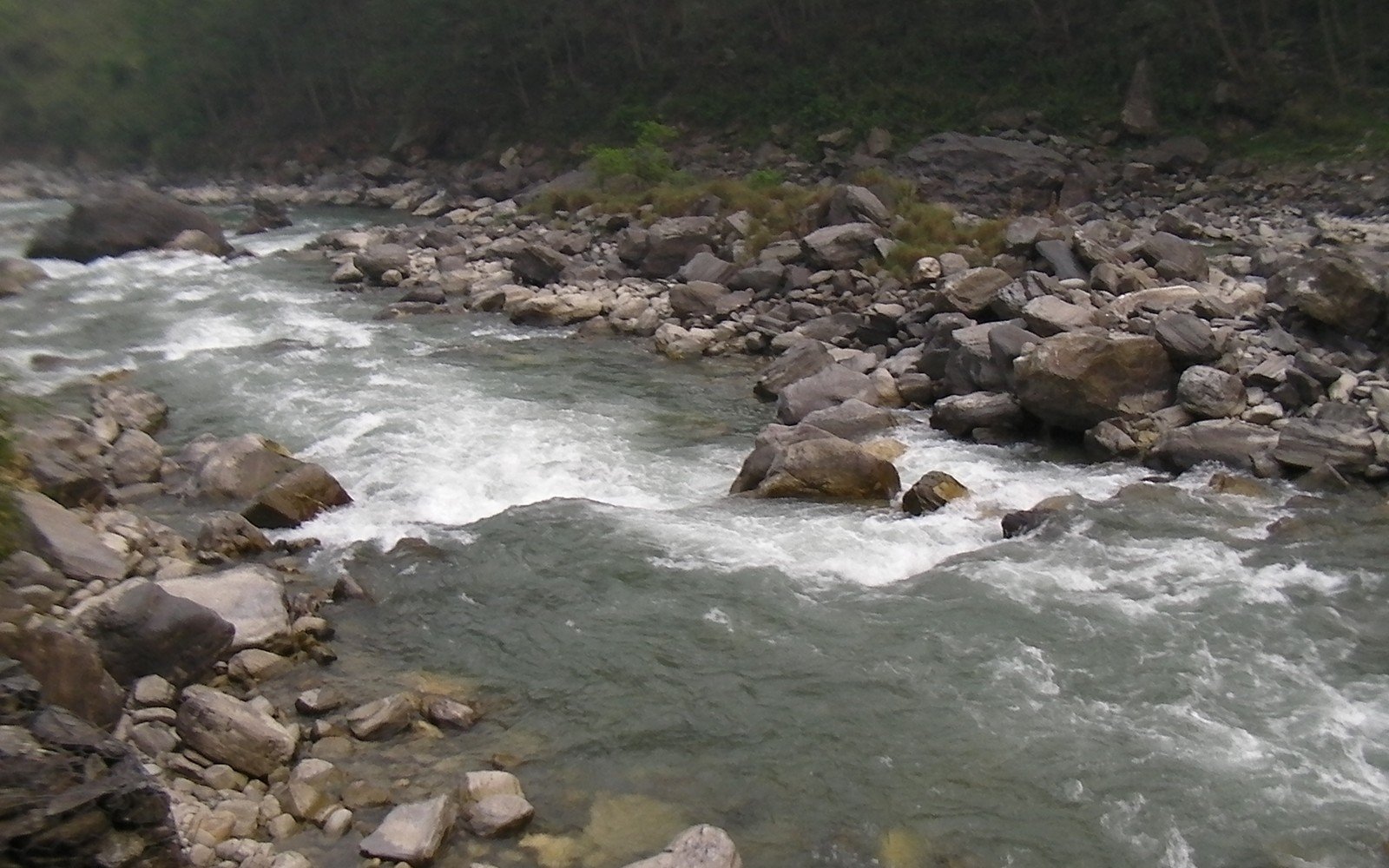 A boulder-strewn Himalayan river running white over rocks between steep forested valley sides in the dry season