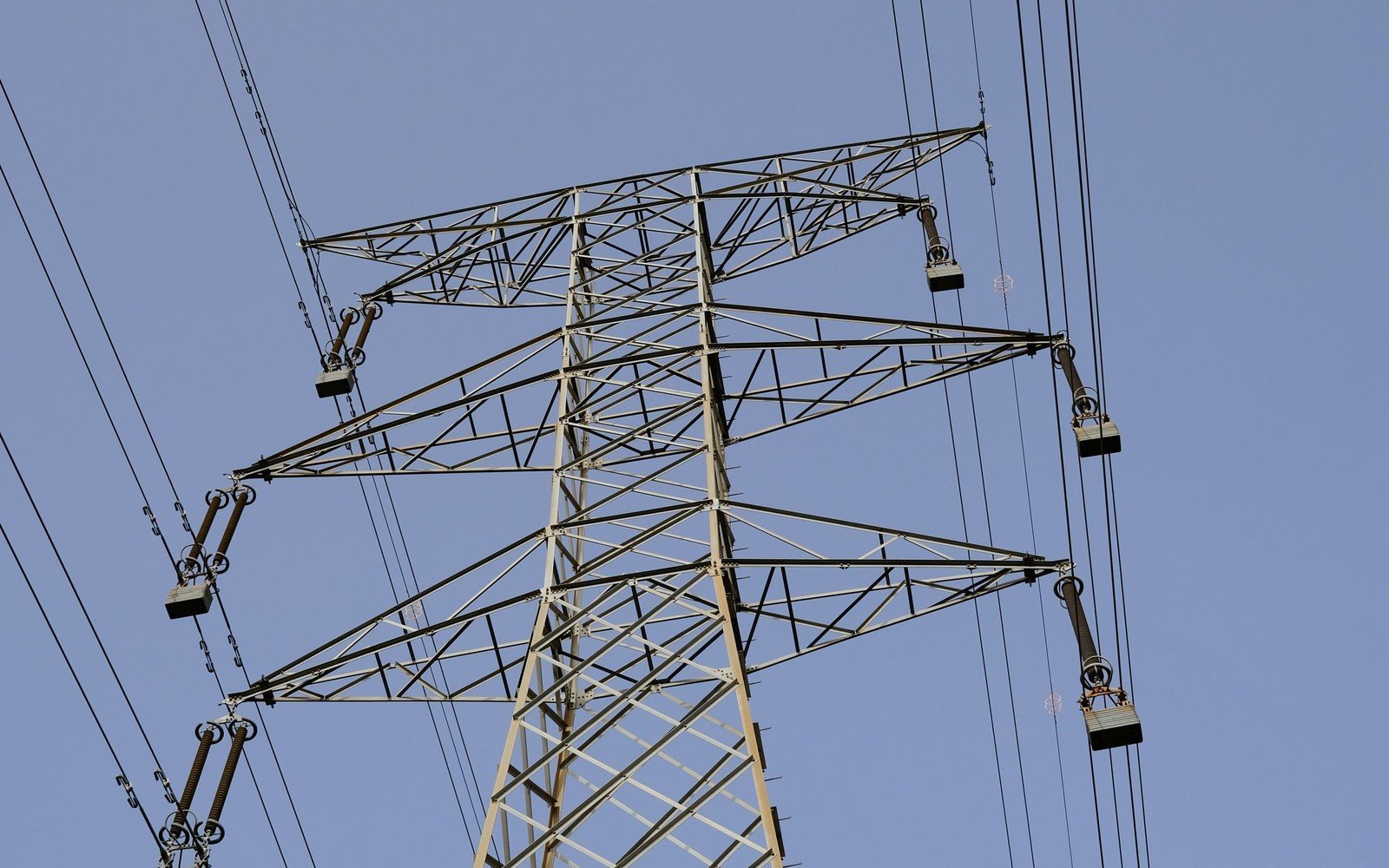 A steel lattice transmission tower photographed from below against a clear blue sky, insulator strings hanging from each crossarm and conductors running out of frame on both sides