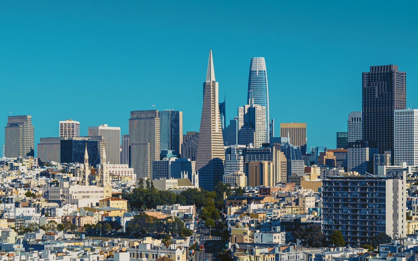 The downtown towers of san francisco seen across low rooftops in clear afternoon light, with a slender pyramid tower and a tapering glass tower standing above the surrounding blocks