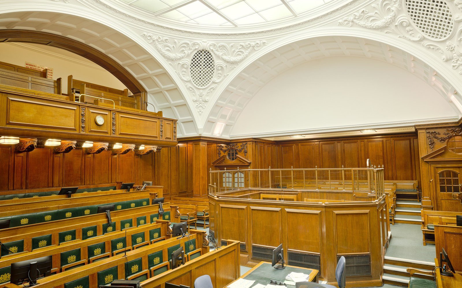 An empty panelled courtroom with oak benches, green leather seats, a glazed enclosure and a coffered barrel ceiling