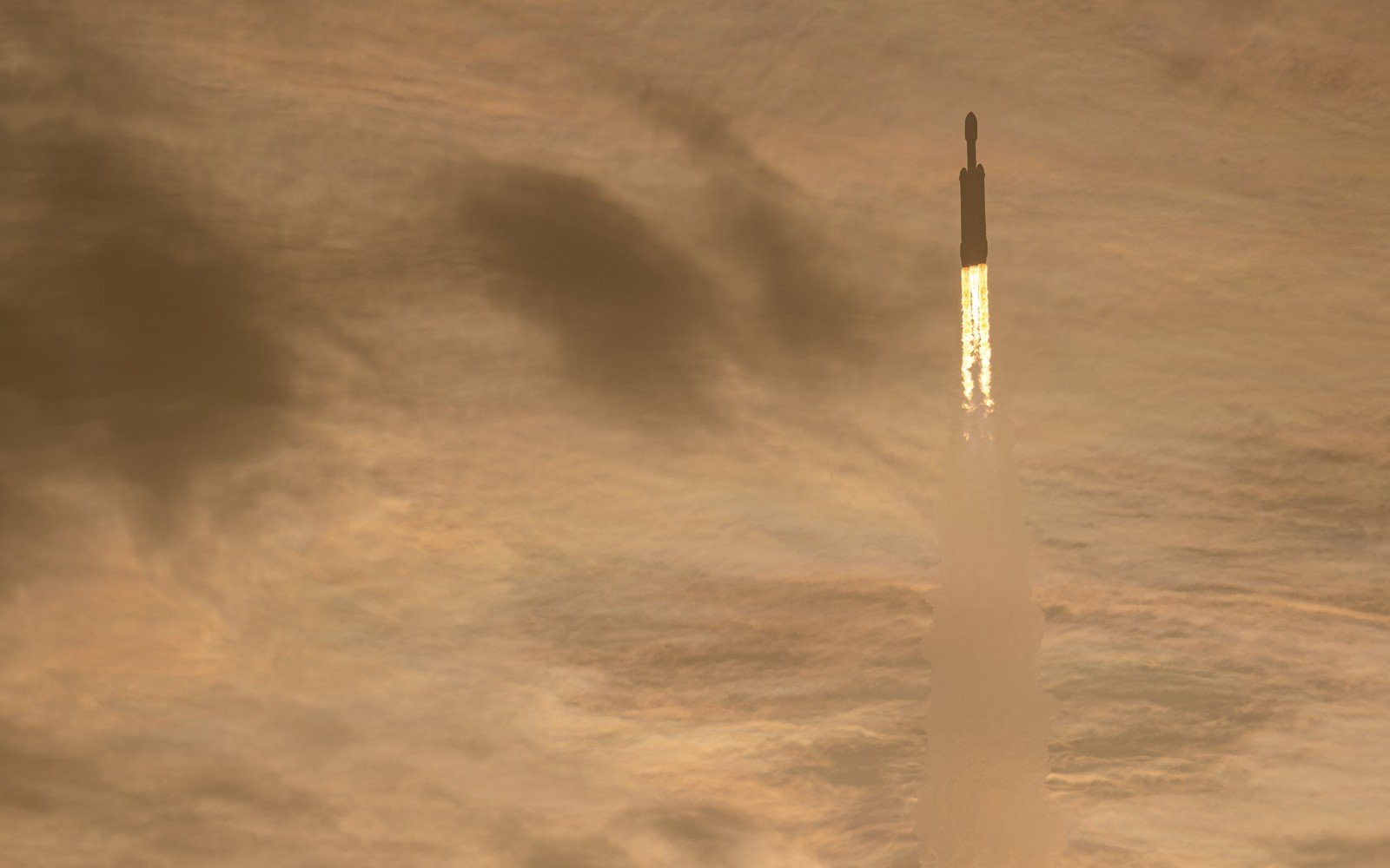 A heavy lift rocket climbing through high cloud on three columns of flame, its exhaust trail lit gold by low sun