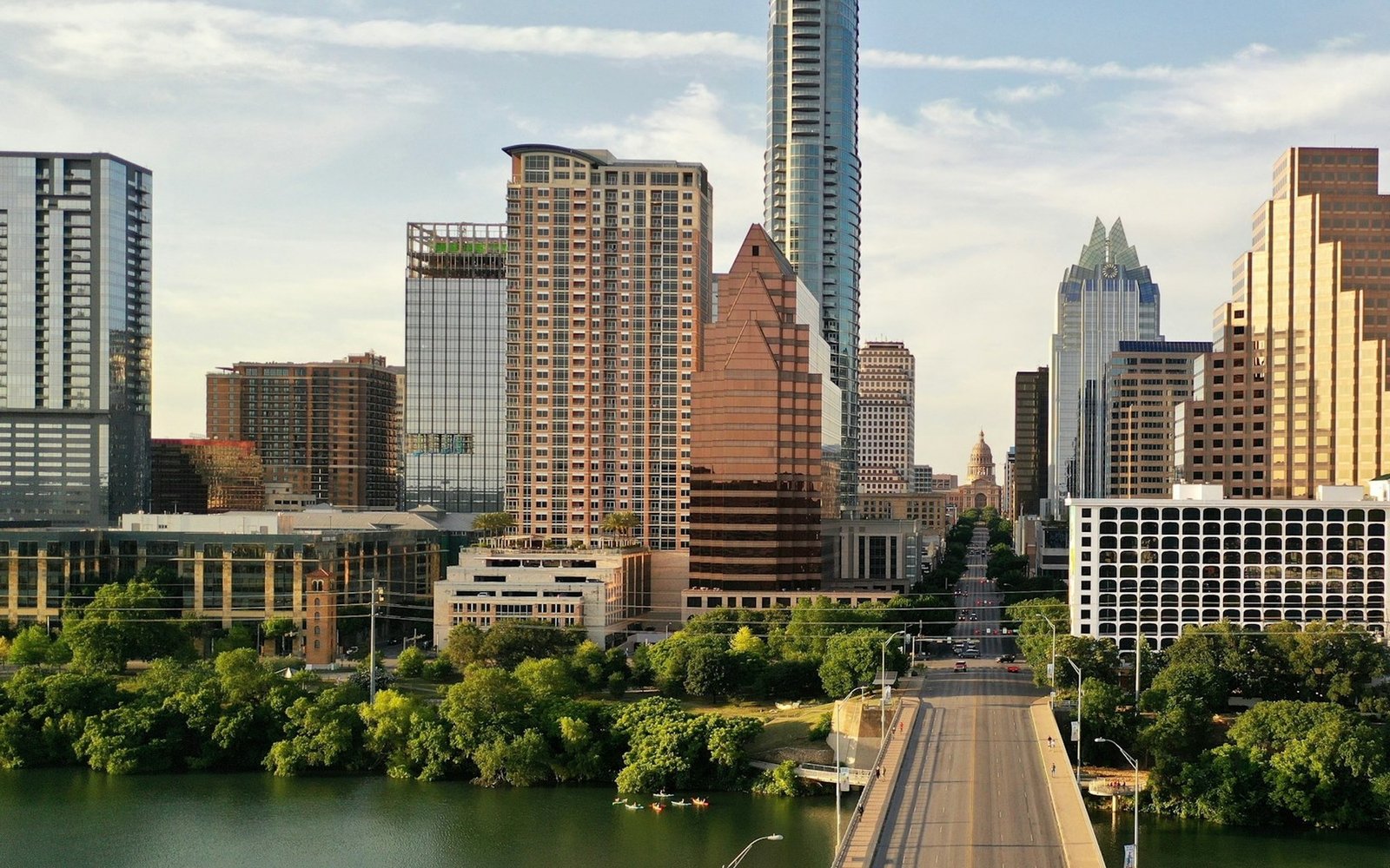 The downtown skyline of Austin, Texas seen from above a bridge crossing a green river, with the state capitol dome visible at the end of a long avenue between the towers