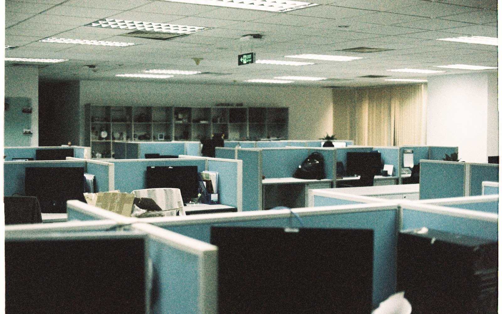 Rows of empty office cubicles with low partitions, dark monitors on each desk and lit ceiling panels above, nobody at any of the workstations