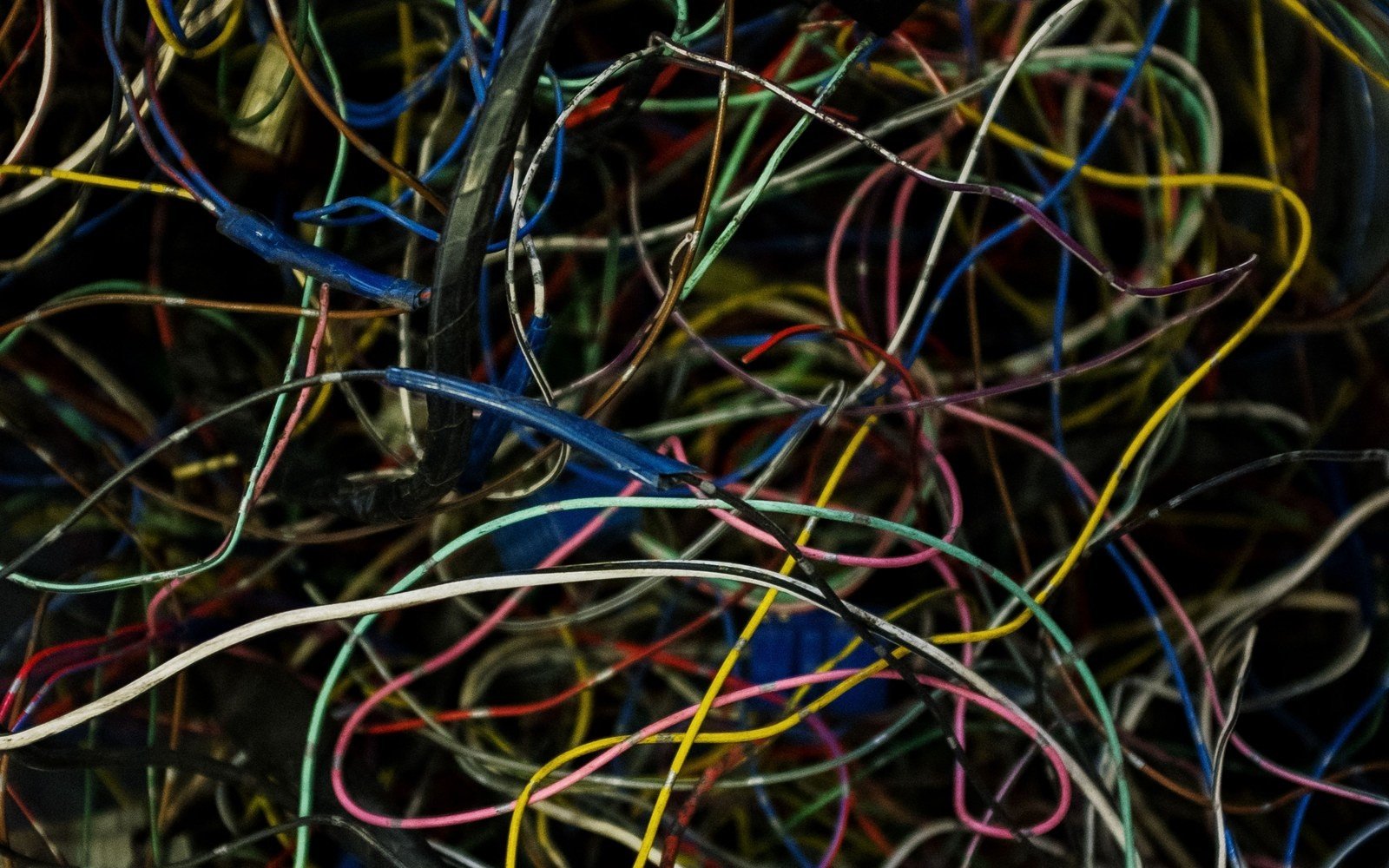 A dense tangle of coloured insulated wires filling the frame, red, blue, yellow, green and white strands crossing over one another in low light