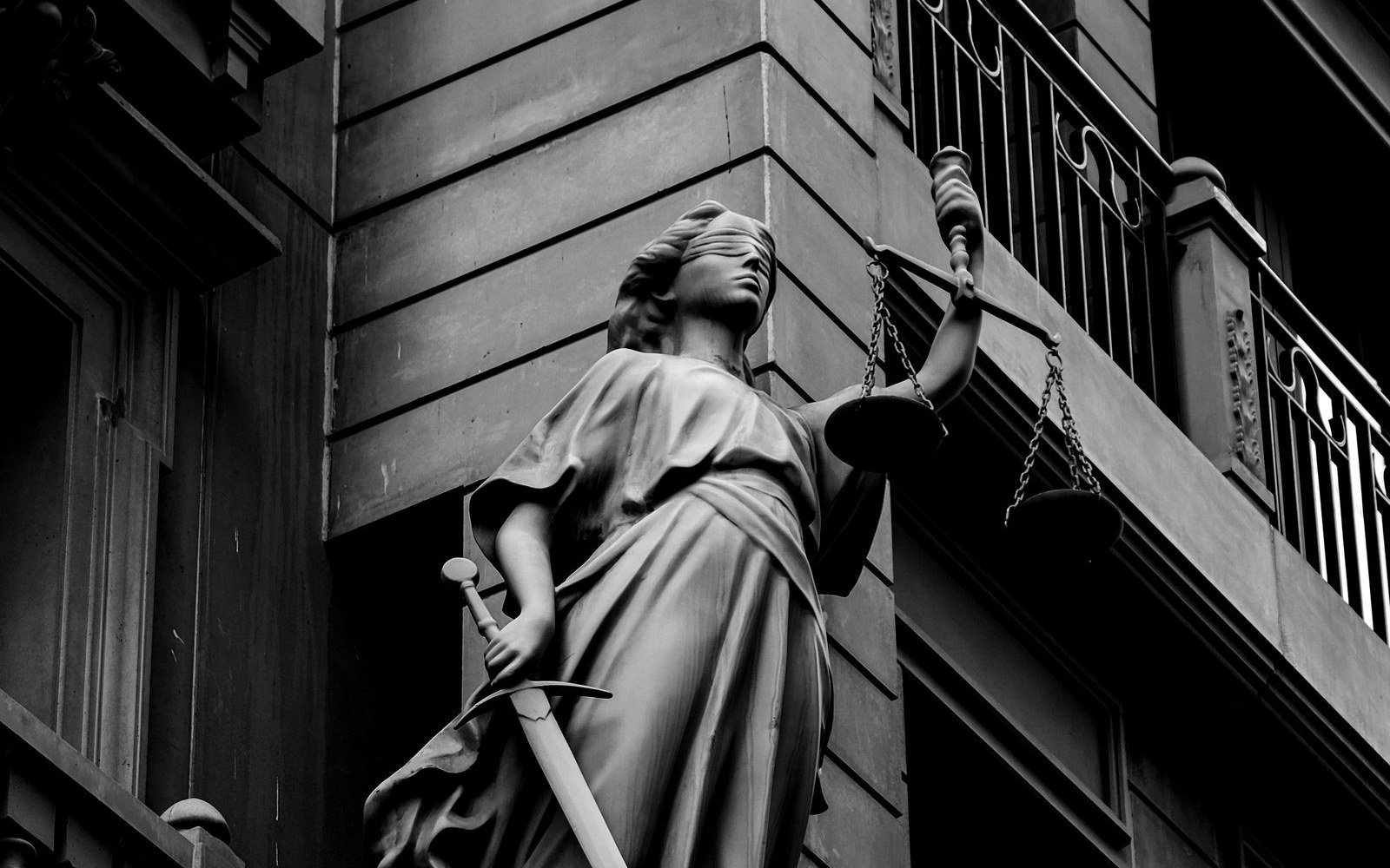 A blindfolded stone figure of Justice holding raised scales in one hand and a downturned sword in the other, against the rusticated stonework of a building, in black and white