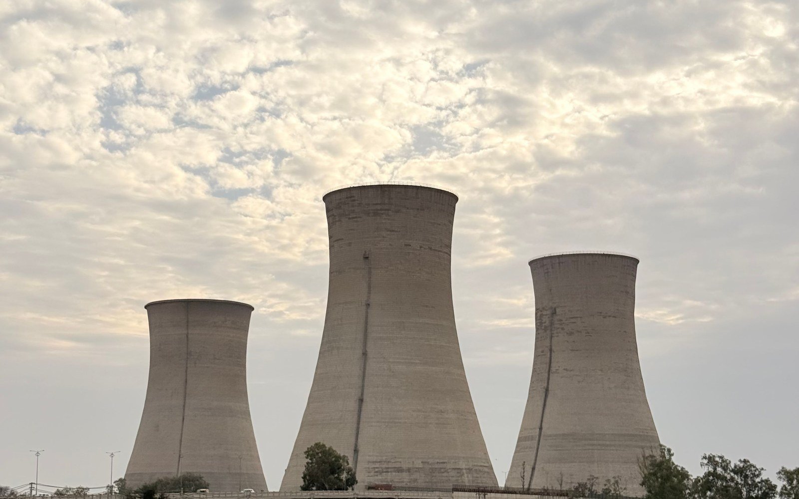 Three concrete hyperbolic cooling towers standing against a pale clouded sky, seen from below with low vegetation along the base of the frame