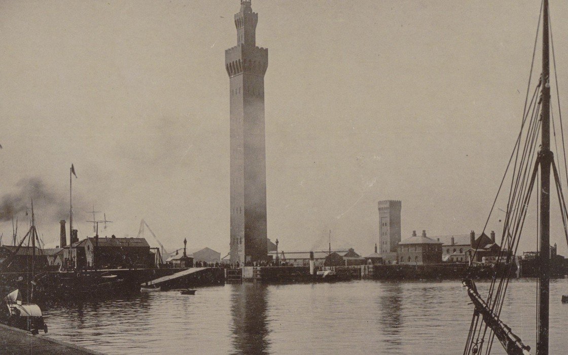 An old photographic print of a tall brick tower standing on a dock quay above still harbour water, with a steam vessel and moored sailing craft alongside