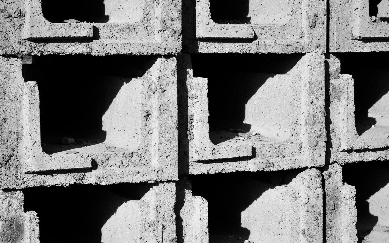 Rows of hollow grey concrete blocks stacked several courses high in hard sunlight, the open cells throwing deep shadows across the rough cast faces