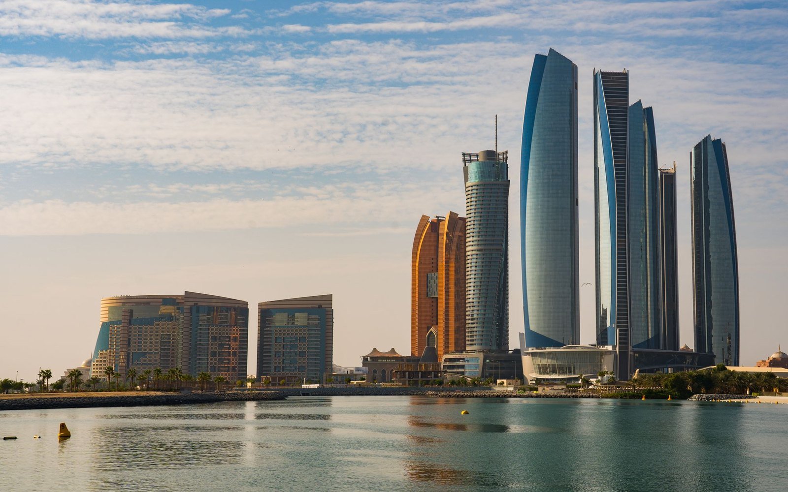 Daylight view across calm water to the Abu Dhabi waterfront, with curved glass towers, mid rise hotel blocks and a low breakwater under a lightly clouded sky