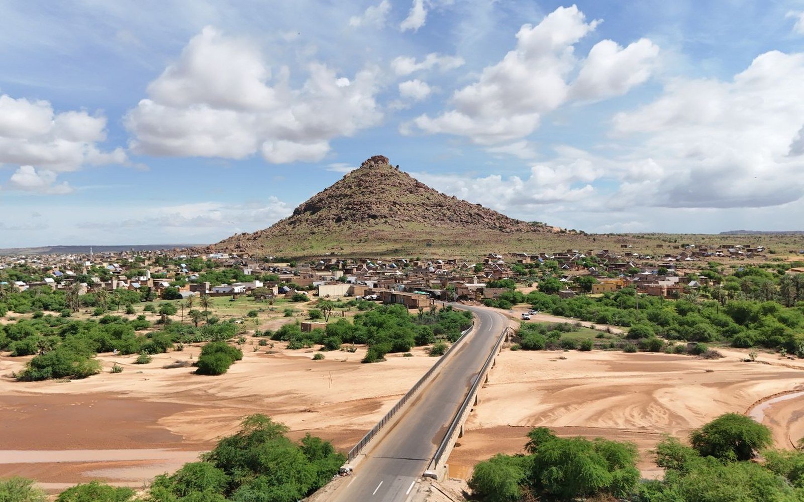 A Malian town of low earth coloured houses spread around a rocky hill, with a road bridge crossing a dry sandy riverbed in the foreground