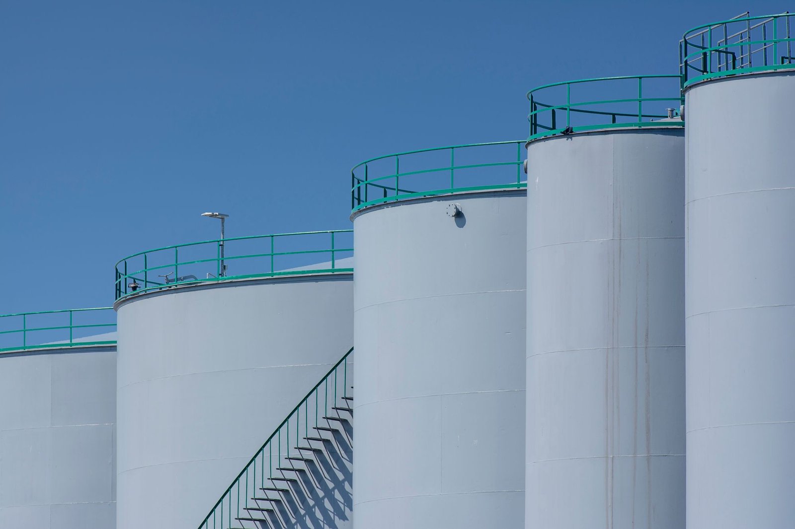 Row of white cylindrical bulk storage tanks with green handrails and an external access stair against a clear sky
