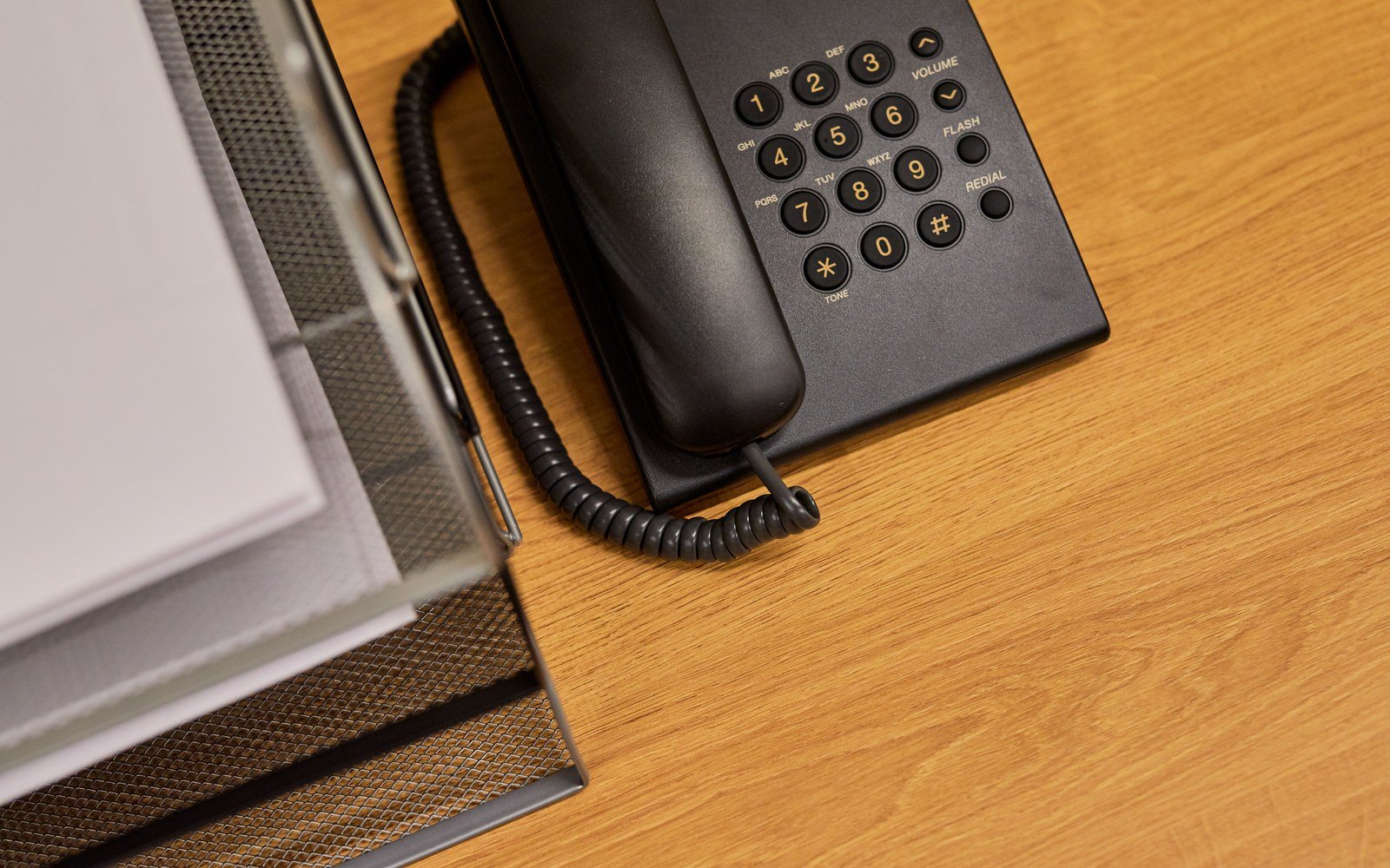 Black push button desk telephone beside a wire document tray on a wooden desk