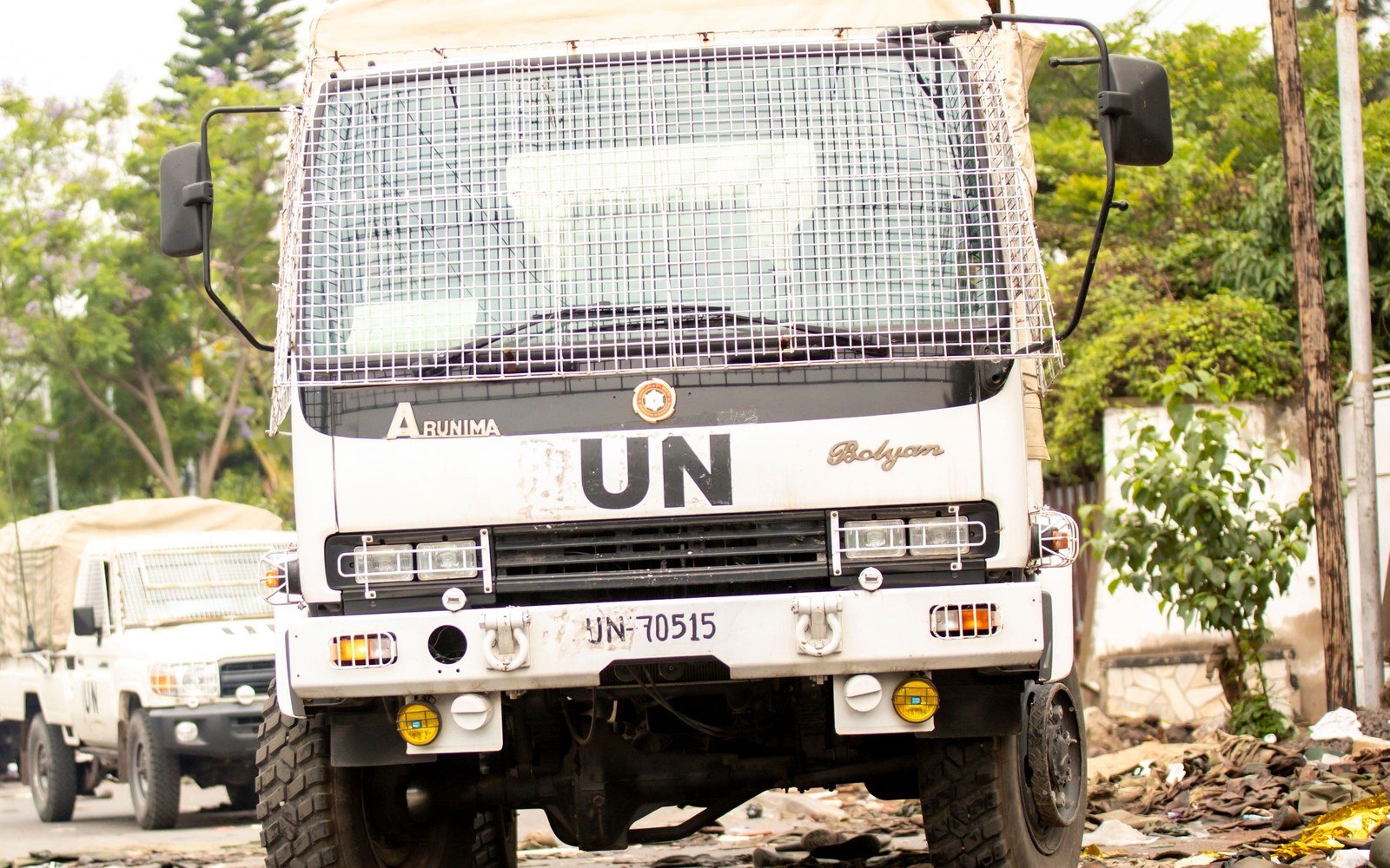 Front view of a white United Nations lorry with a mesh windscreen guard standing on a road strewn with debris, a second white vehicle behind it and trees along the roadside