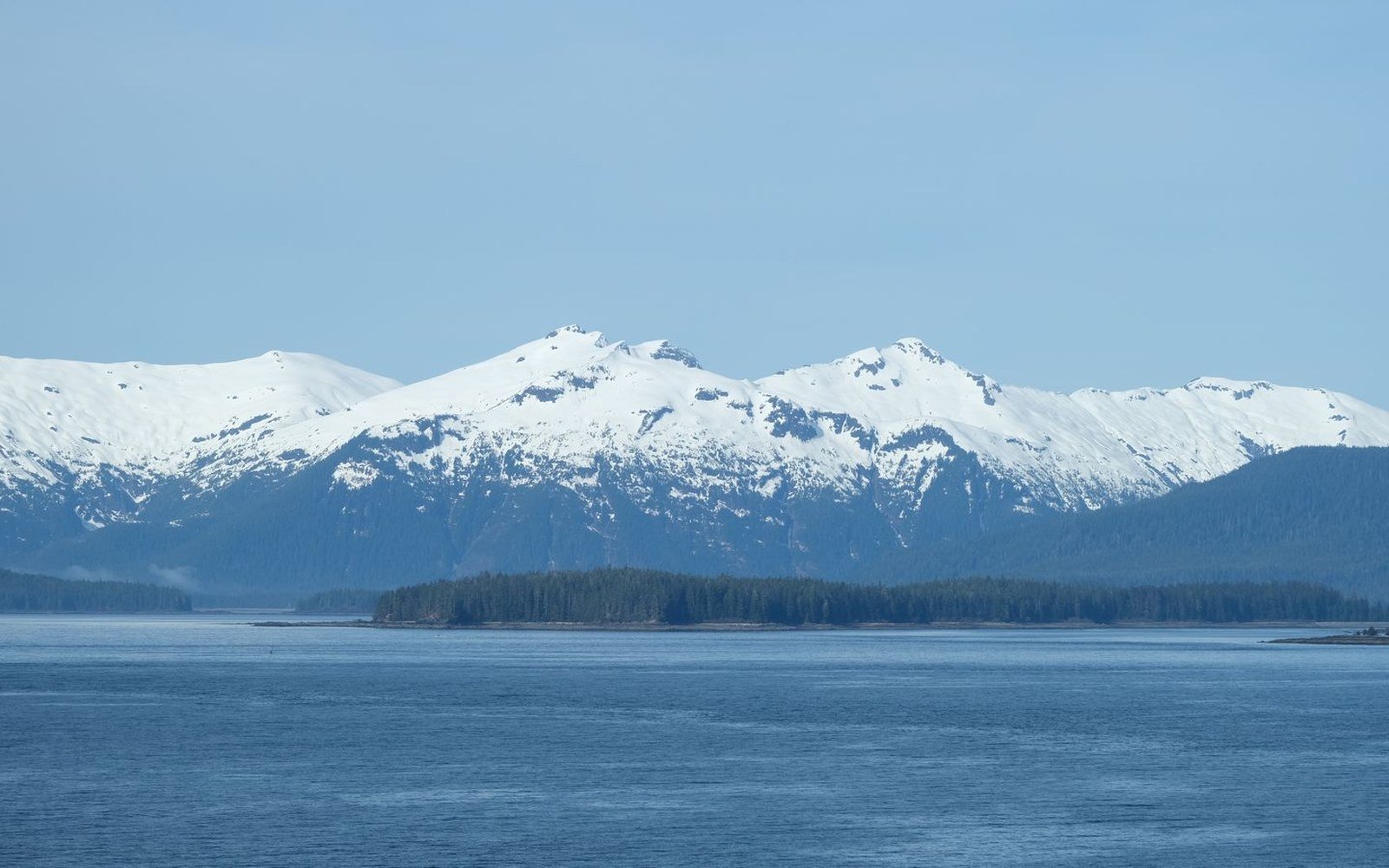 Snow topped mountains above a wooded shoreline and open water in Alaska, seen from the sea on a clear day