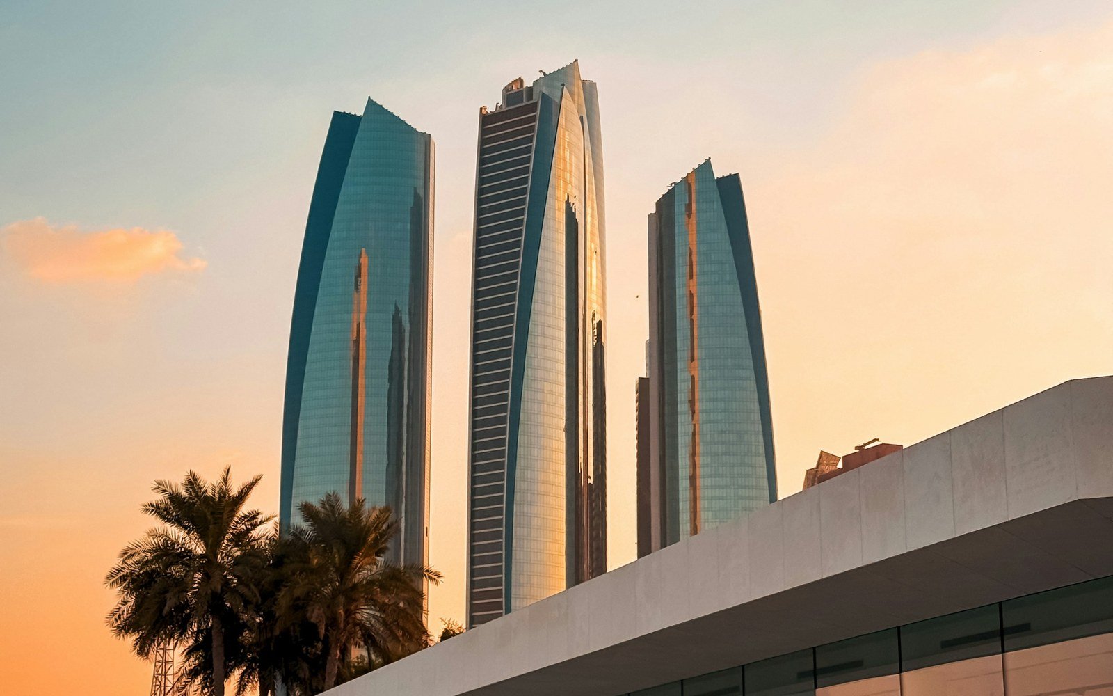 Three tapered glass towers catching a warm orange sky above the pale roof line of a low building, with a palm frond at the left edge of the frame