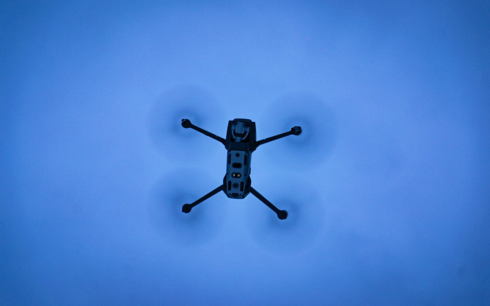 The underside of a small four-rotor drone in flight against a blue sky, its camera and sensors visible