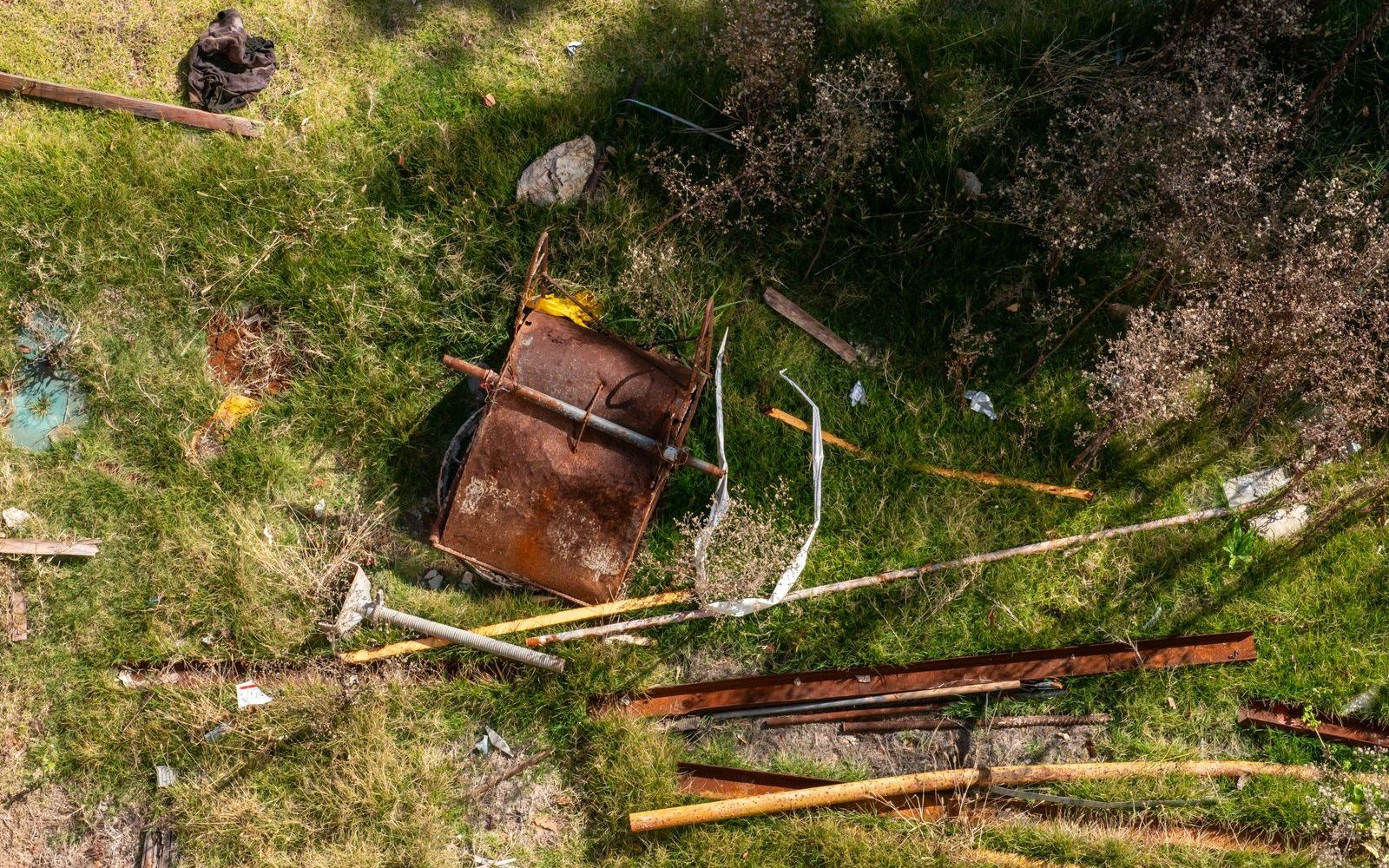 Rusting scrap metal, lengths of timber and an overturned barrow lying dumped in rough grass, seen from above