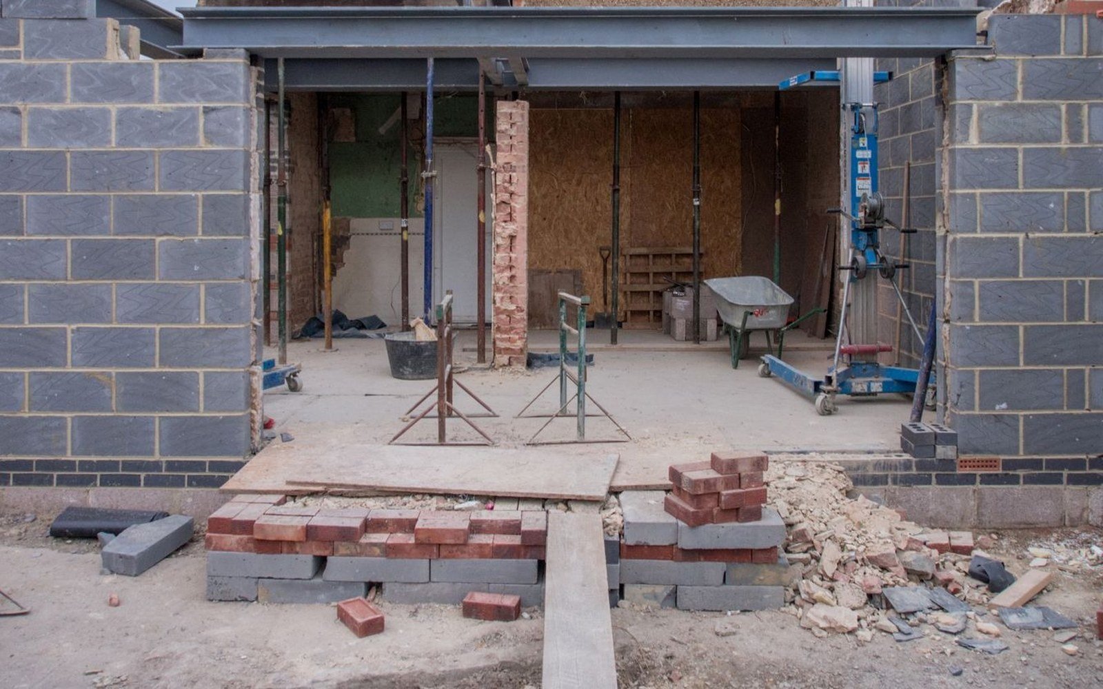 A rear house extension under construction, with a steel beam over the opening, adjustable props beneath it and stacks of bricks and blocks on the ground
