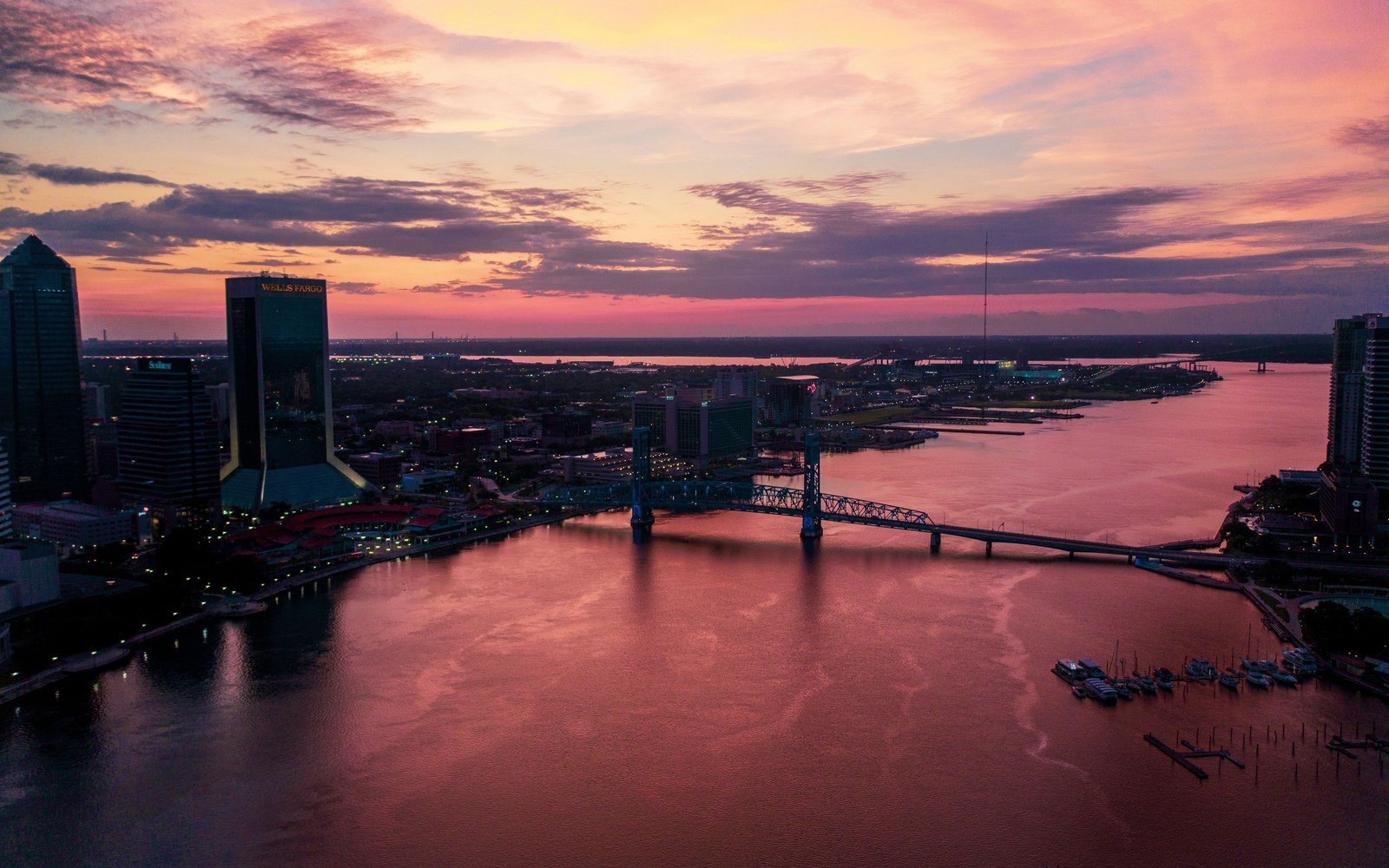 The Jacksonville skyline at sunset seen across the St Johns River, office towers on the left and a lift bridge crossing water lit pink by the sky