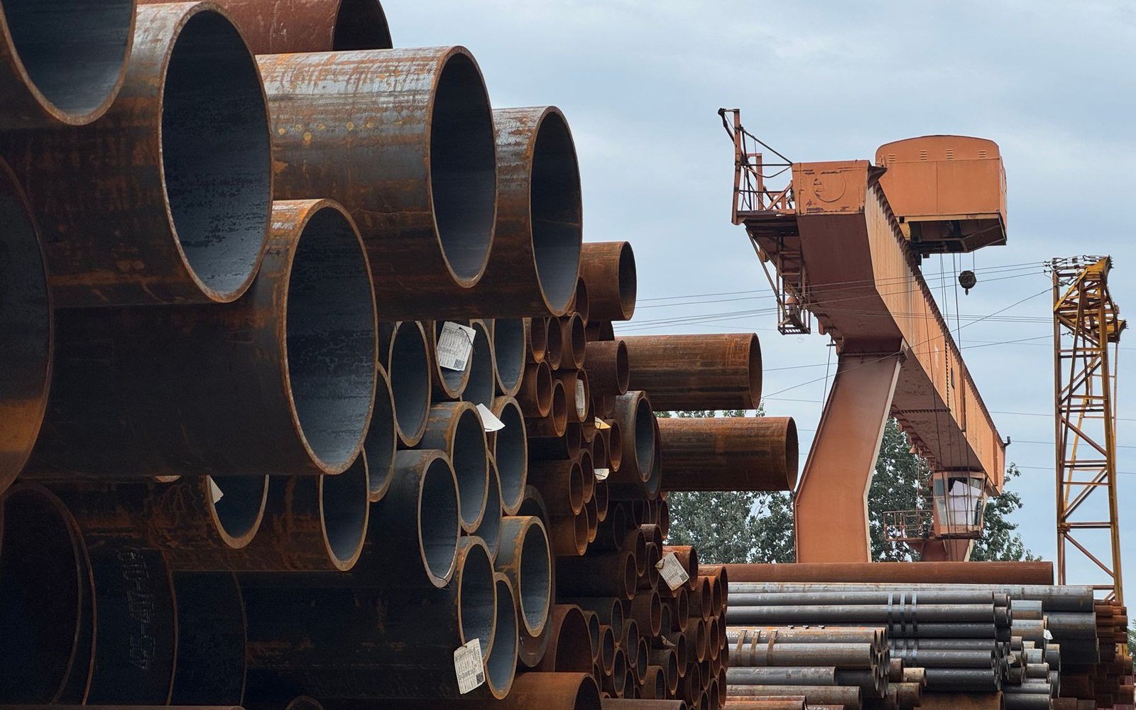 Stacked lengths of dark steel tube of several diameters in an open storage yard, a gantry crane behind them