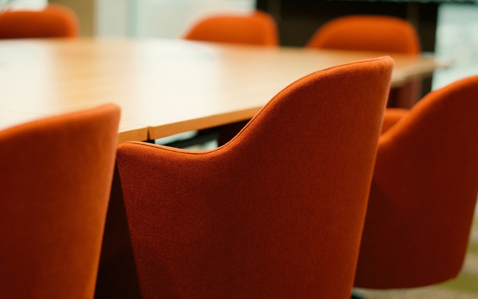 An empty boardroom with a long pale wood table and a row of high backed rust coloured upholstered chairs, shot low and close along the table edge