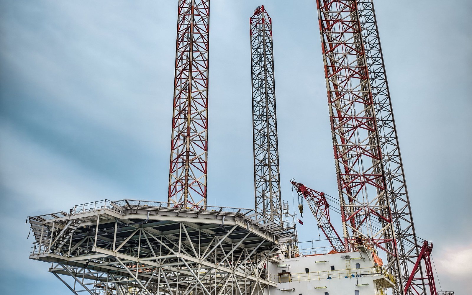 Close daylight view of a jack-up drilling rig, showing three latticed legs, the cantilever deck and the rig crane against a pale clouded sky