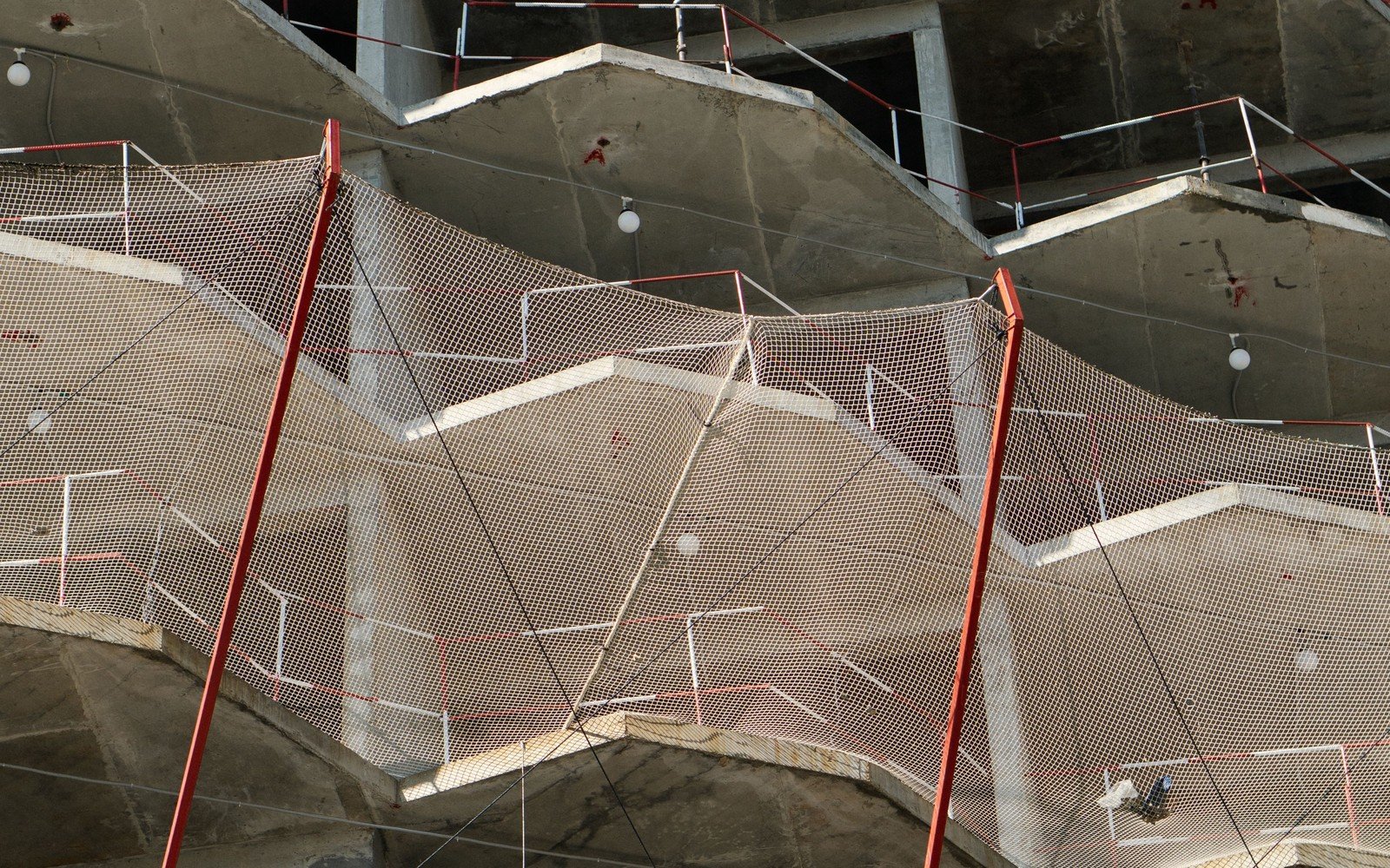 Cast concrete balcony slabs on a residential frame under construction, edge protection netting strung between red posts across three storeys, with temporary lighting hung from the soffits