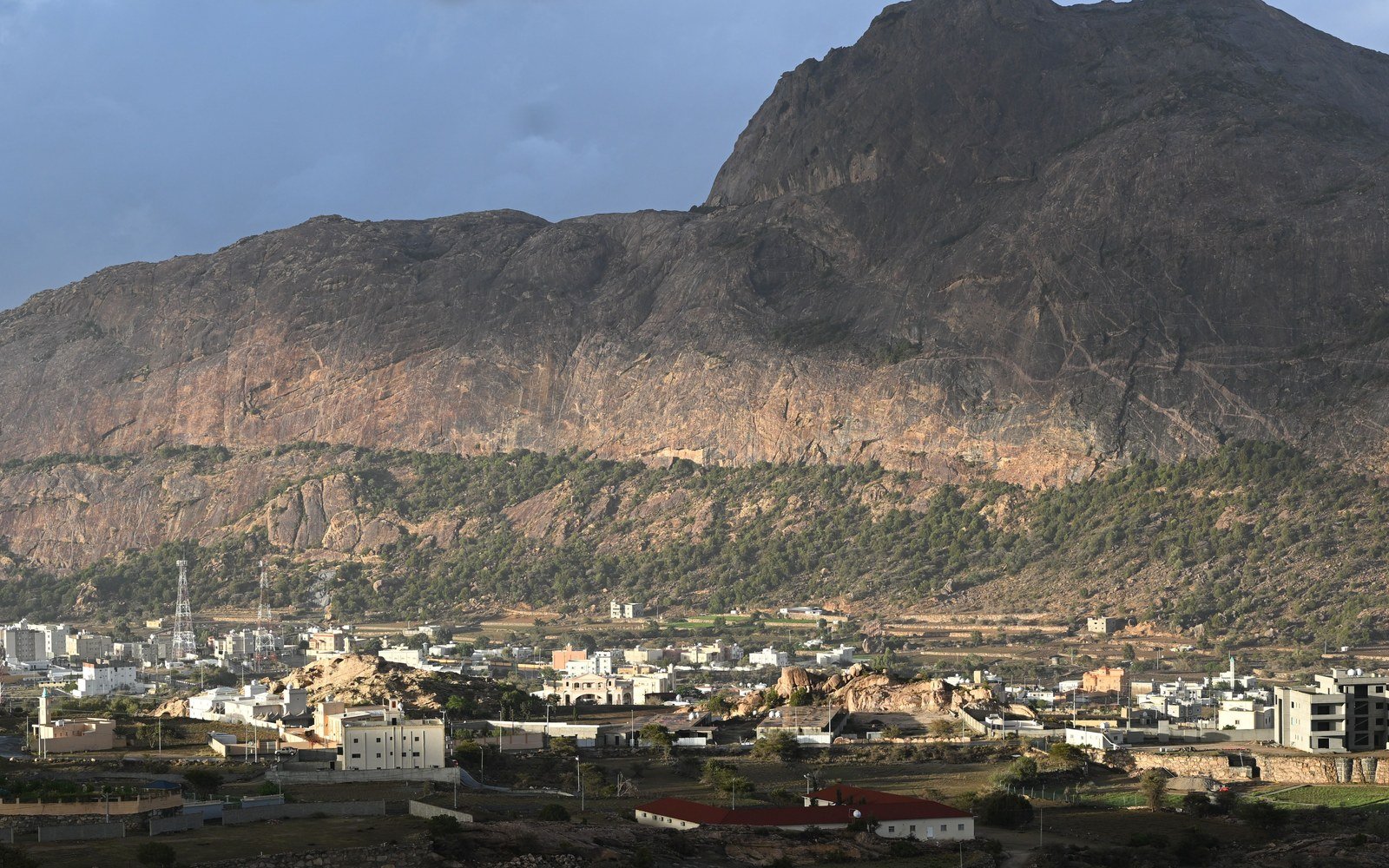 A bare rock massif of the Arabian Shield rising in late afternoon light above a low town of white and sand coloured buildings, with exposed granite outcrops in the foreground