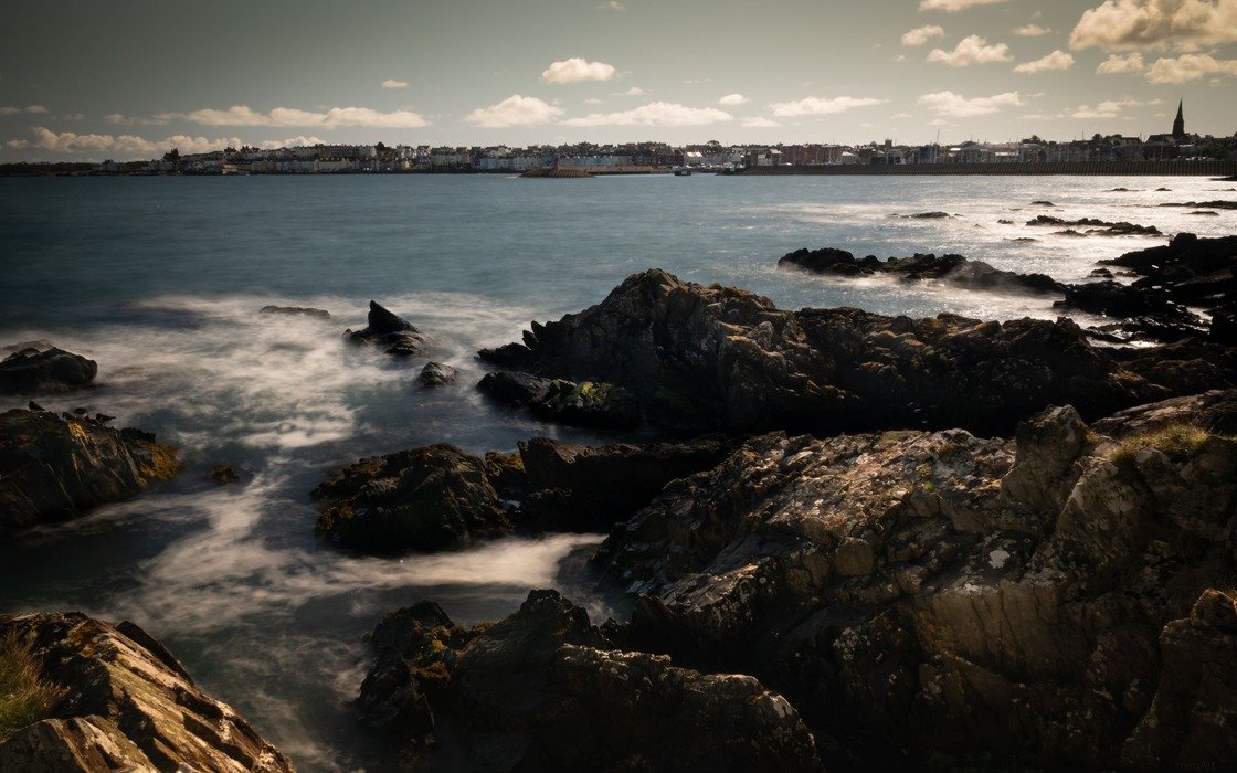 A rocky foreshore in Bangor with the sea washing between the rocks and the low roofline of the town along the far shore of the bay