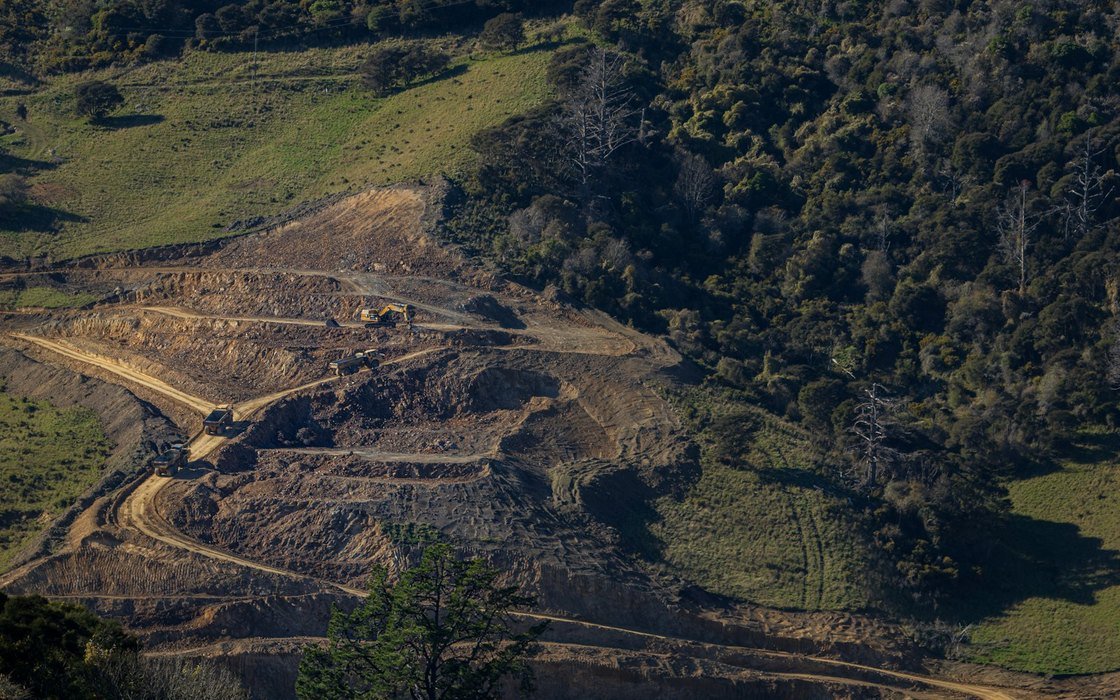 A tracked excavator and articulated dump trucks working benched earthworks cut into a wooded hillside, with haul roads between the benches