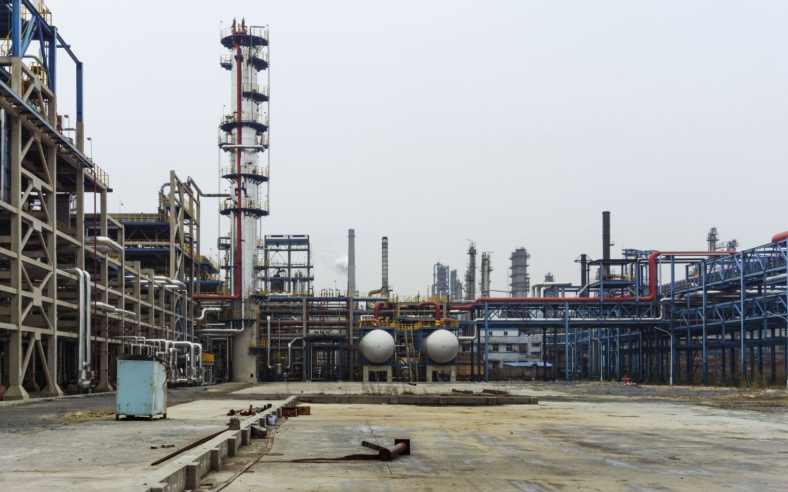 A petrochemical process plant seen across an empty concrete hardstanding, steel pipe racks and a lattice framed column in the middle distance, two white spherical storage vessels at ground level and a flat pale sky above