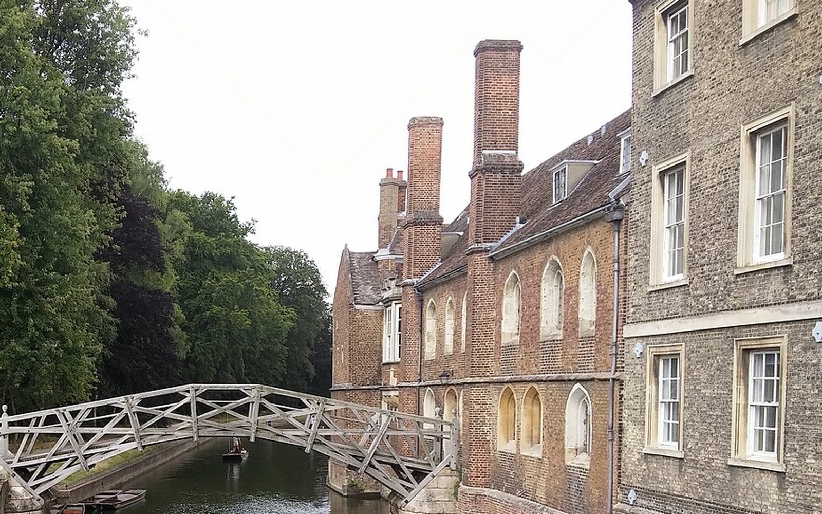 A white timber footbridge arching over a narrow river beside an old brick and stone college range in Cambridge