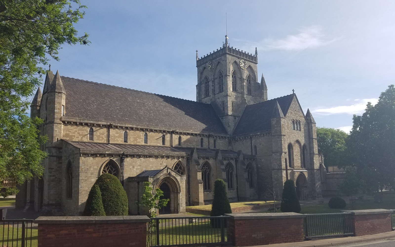 A large medieval church with a square crossing tower behind railings, seen from the road across mown grass