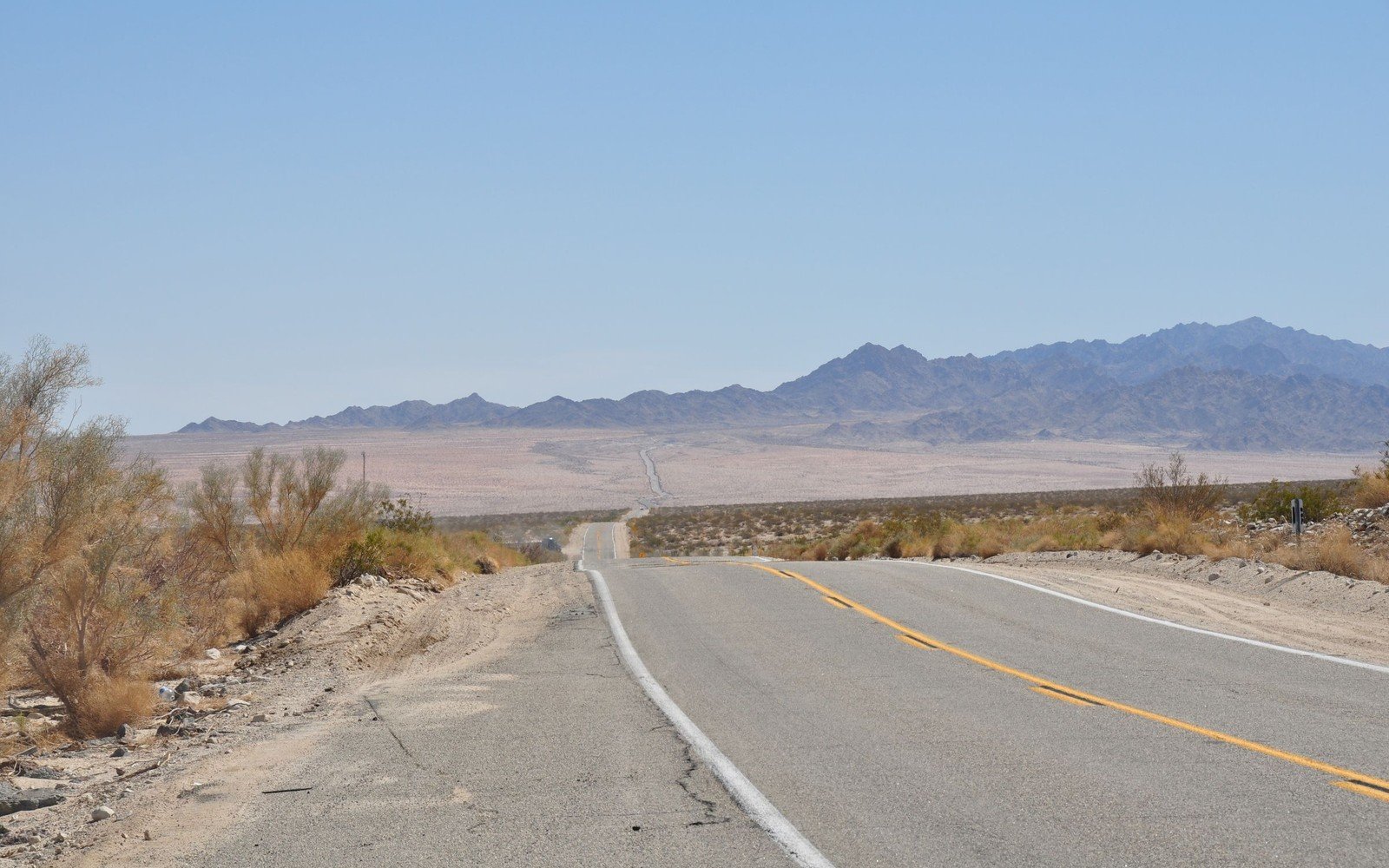 An empty two lane asphalt road running straight across the Californian desert towards a bare mountain range under a clear sky