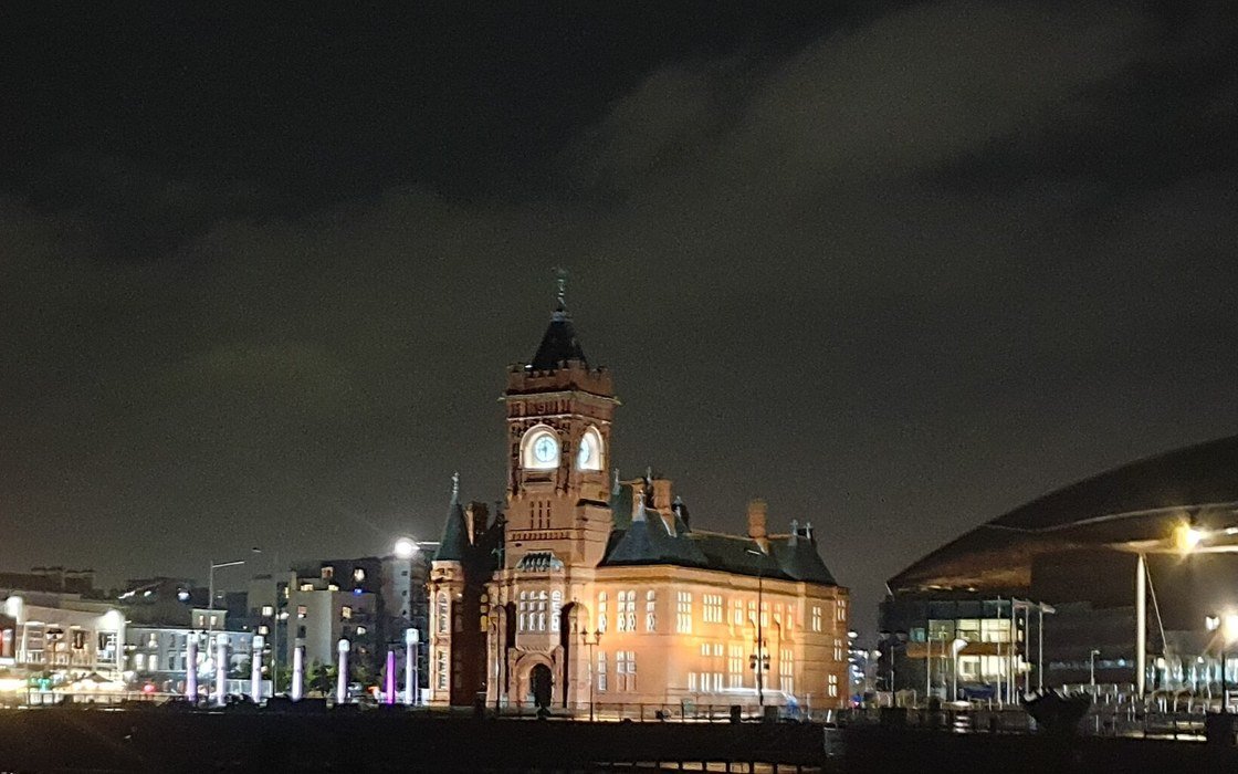 A lit Victorian clock tower and waterfront buildings reflected in dark water at night