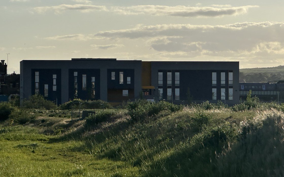 A modern dark clad English school building seen across rough grass in evening light, with no people in frame
