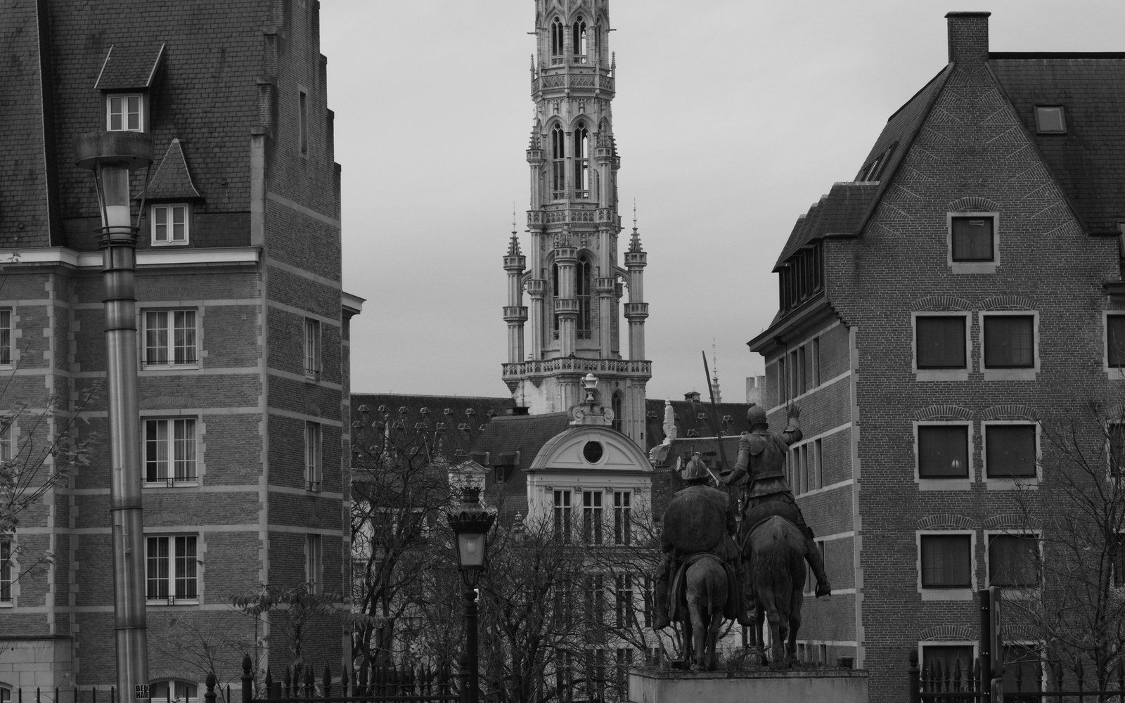 A monochrome view along a Belgian street towards a tall gothic civic tower, with stepped gable roofs and a bronze equestrian statue in the middle distance
