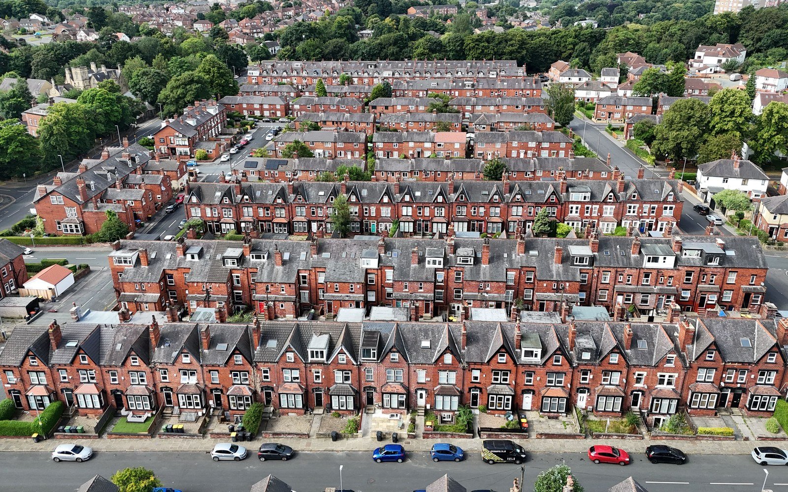 An elevated view of long rows of red brick terraced houses with bay windows and slate roofs, with cars parked along the street below