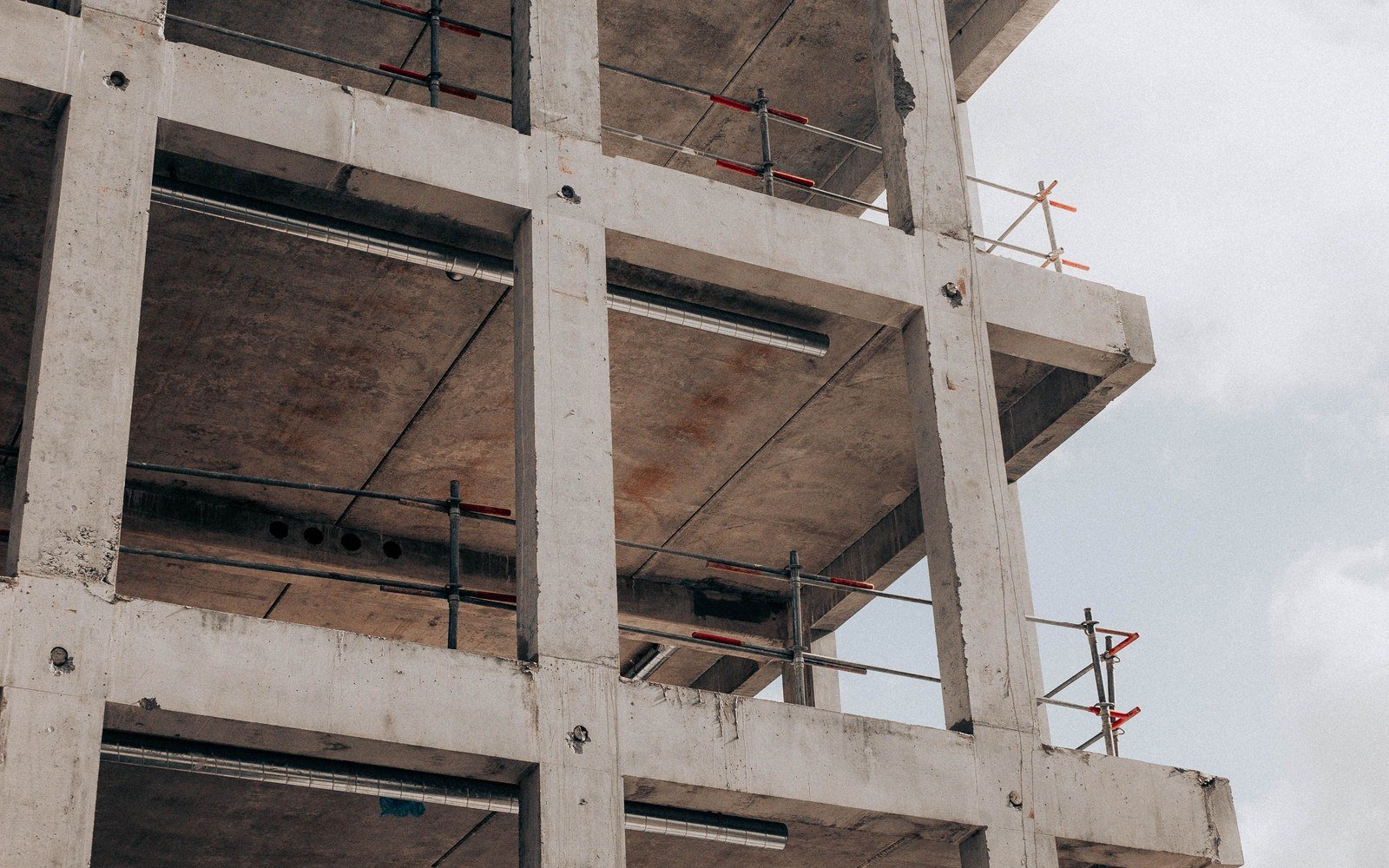 The unfinished reinforced concrete frame of a multi storey building seen from below, columns and downstand beams exposed and a red and white prop against the soffit