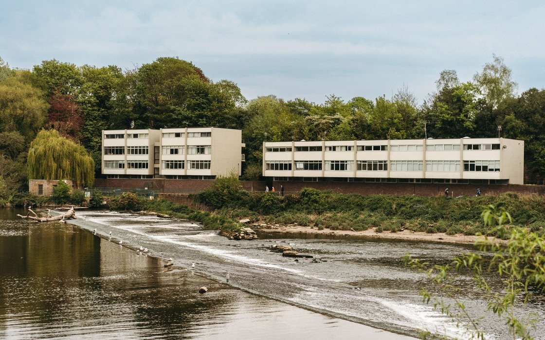 A weir across a wide river in Chester with modern white riverside buildings and mature trees on the far bank