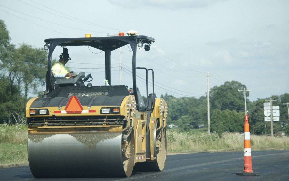 A road roller compacting a carriageway beside a traffic cone on a summer day