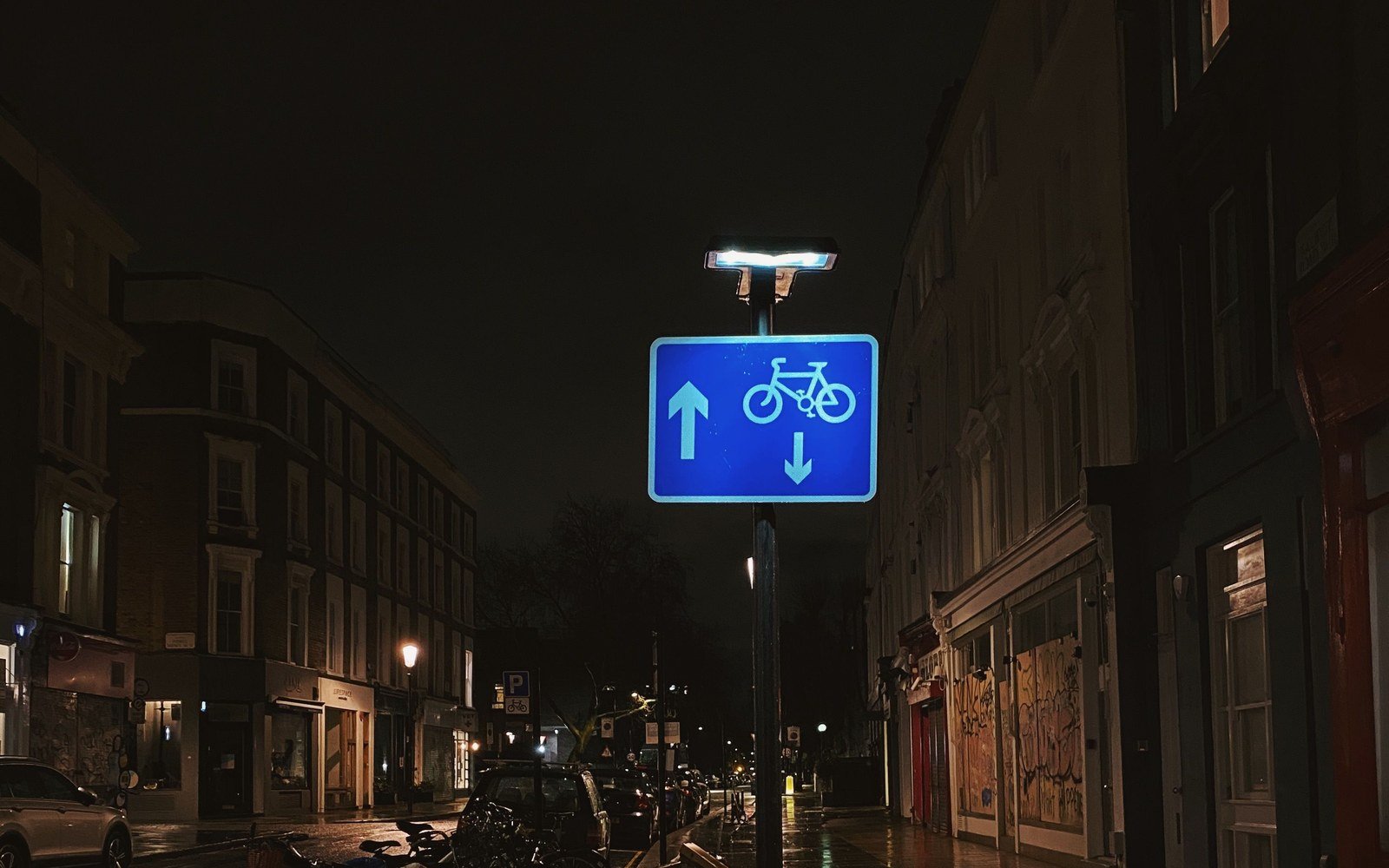 A wet city street at night with a blue cycle route sign on a lamp column and reflections on the road surface