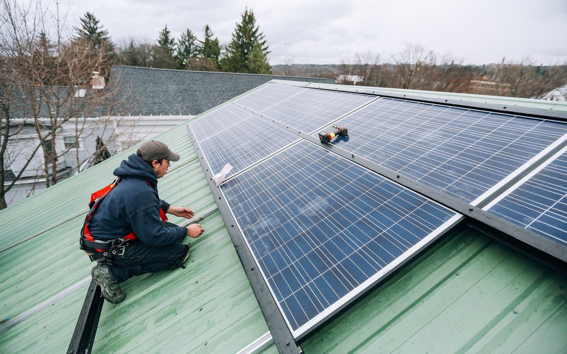 A technician kneeling on a pitched metal roof while fixing a solar photovoltaic panel into its rail