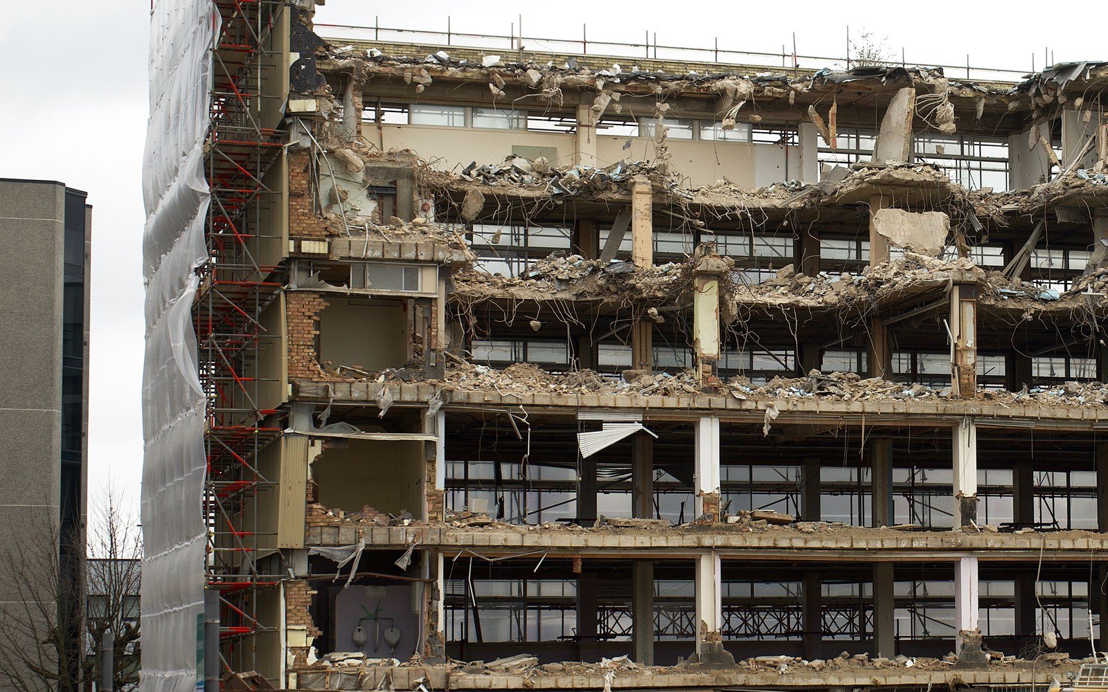 A multi-storey concrete framed building part way through being taken down, with floor slabs exposed, rubble on the ground and scaffolding and protective sheeting to one side