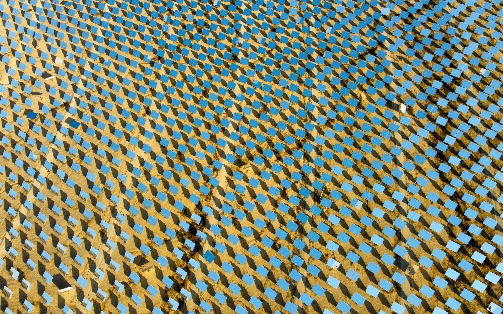 Daylight aerial view across a dense field of angled solar mirrors laid in regular rows over pale sandy ground, with no buildings, vehicles or people in the frame