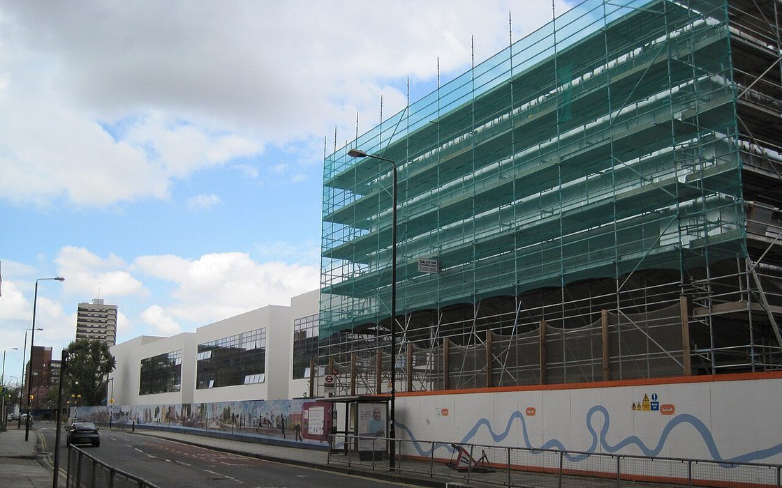 A school building under construction behind green netted scaffolding, with site hoarding along the pavement