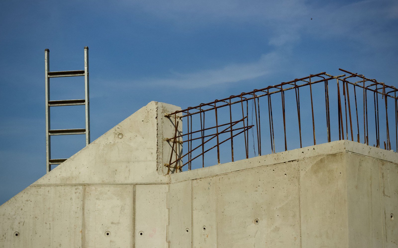 The top of an unfinished reinforced concrete wall against a blue sky, starter bars projecting from the slab edge and tied into a line of horizontal steel, a shuttering panel still fixed below the bars and a metal ladder standing clear of the wall on the left