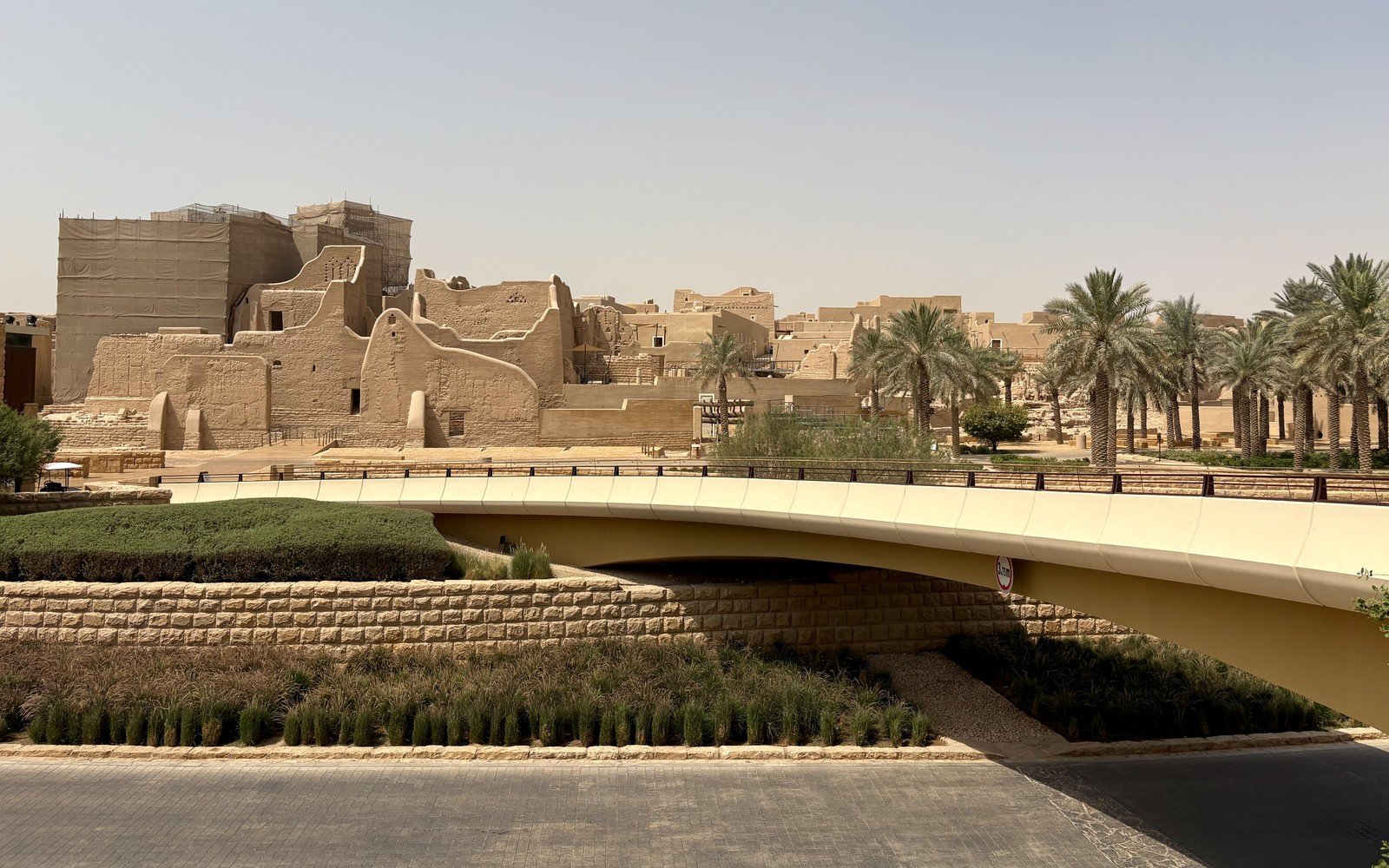 Restored and part restored mudbrick buildings of the At-Turaif district in Diriyah stand behind date palms and a modern pedestrian bridge in daylight