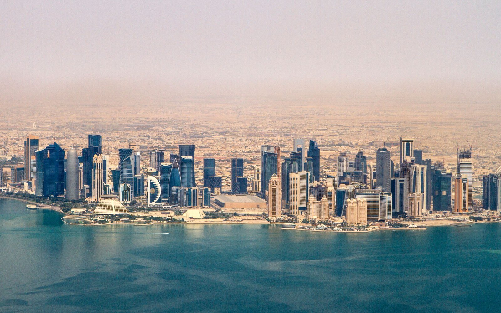 An aerial view of the Doha waterfront on a clear day, with the towers of the business district on a headland above a bay and low-rise development spreading inland behind them